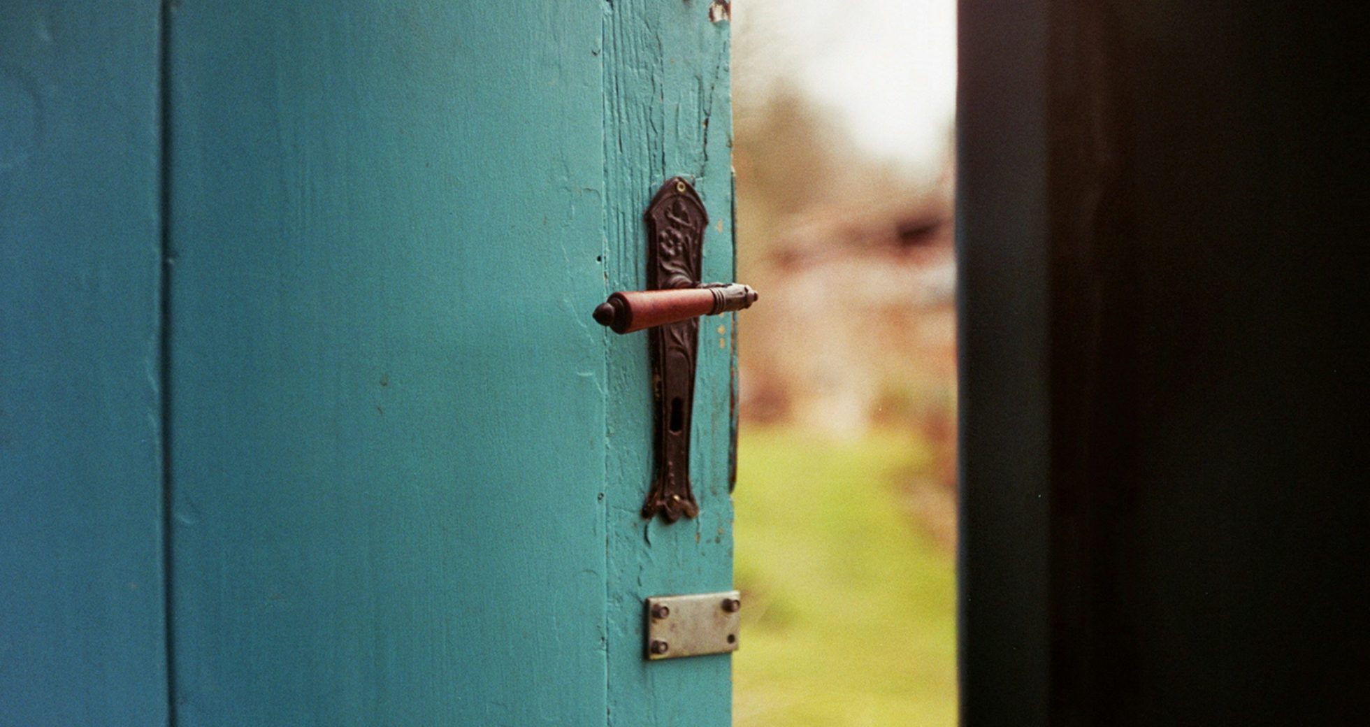 A weathered teal wooden door with a clay-red handle, half open onto a sunlit garden